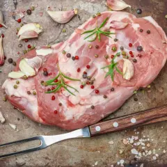 Surowe mięso jagnięce, czosnek, rozmaryn i kolorowy pieprz na blaszce, gotowe do peklowania na mokro. Widelec obok.