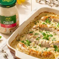 Pyszna kiełbasa w sosie chrzanowym, zapiekana w naczyniu, z dodatkiem ziół. Obok słoik sosu chrzanowego.