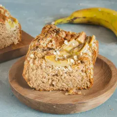 Kawałek pysznego, domowego chlebka bananowego fit, posypanego płatkami owsianymi. Obok leży dojrzały banan.