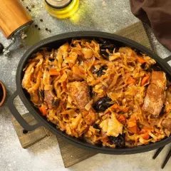 Pyszny bigos z kiszonej kapusty z mięsem i śliwkami, podany w żeliwnej patelni. Obok miseczka ze śliwkami i młynek do pieprzu.