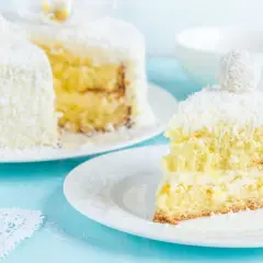Pyszne ciasto rafaello na biszkopcie, obsypane wiórkami kokosowymi. Kawałek na talerzyku kusi kremowym nadzieniem.