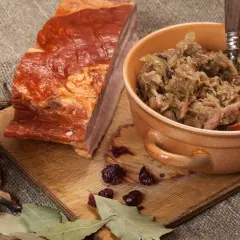 Ania gotuje bigos. Na desce kawałek wędzonego boczku, miseczka z bigosem, kieliszek czerwonego wina, suszone grzyby i liście laurowe.