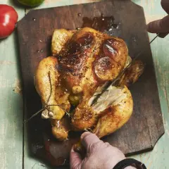 Złocisty kurczak z rożna, pieczony do perfekcji, kroimy na drewnianej desce. Obok leżą pomidor i zielona papryka.