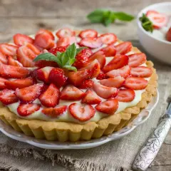 Pyszna tarta z truskawkami, udekorowana listkami mięty, na drewnianym blacie. Obok miseczka z deserem i nóż.
