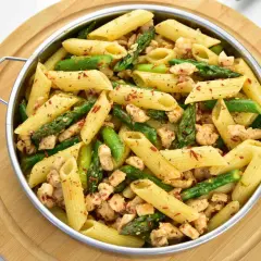 Penne z kurczakiem i szparagami, posypane płatkami chili, w metalowej misce na drewnianej desce.