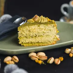 Pyszne ciasto z kremem pistacjowym, ozdobione orzechami i podane na talerzu. Obok filiżanka kawy i łyżeczka.