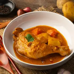 Pyszny kurczak w sosie curry z ziemniakami, podany w białej misce na lnianym tle. Obok widoczne przyprawy i drewniane sztućce.