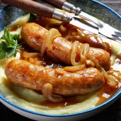 Złocista kiełbasa z cebulą i kremowym purée ziemniaczanym, udekorowana natką pietruszki. Danie podane w niebieskiej misce, gotowe do jedzenia.