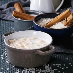 Gotowy do podania ryż na mleku w ceramicznej miseczce, obok miseczki z ryżem i laskami cynamonu.