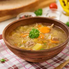 Gęsty, aromatyczny kapuśniak z kiszonej kapusty, z kawałkami mięsa i warzyw, podany w glinianej misce na kratkowanej serwetce.