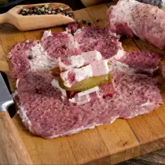 Roladki z karkówki w trakcie przygotowania. Na desce leżą rozbite plastry mięsa, doprawione pieprzem i solą, z dodatkiem boczku i ogórka kiszonego. Obok nóż i drewniana łyżka z ziarnami pieprzu.