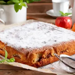 Puszysty biszkopt z jabłkami, posypany cukrem pudrem, obok jabłka i cynamon.