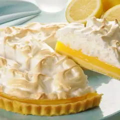Puszysta tarta cytrynowa z bezą, z wyciętym kawałkiem, prezentuje się apetycznie. Obok leżą świeże cytryny.