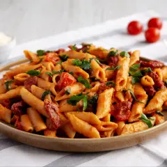 Penne z sosem pomidorowym i suszonymi pomidorami, posypane świeżą natką pietruszki. Obok miseczka z parmezanem i kilka pomidorków koktajlowych.