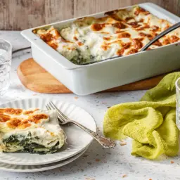 Pyszne lasagne ze szpinakiem, zapieczone na złoto, podane na talerzu obok szklanki wody z cytryną.