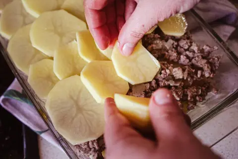 Ręce układają plastry ziemniaków na warstwie mięsa mielonego, tworząc pyszną zapiekankę ziemniaczaną z mięsem mielonym.