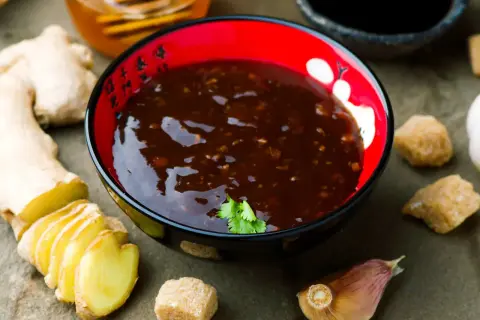 Sos teriyaki przepis: miseczka z ciemnym sosem, obok plasterki imbiru, czosnek, cukier i miód.