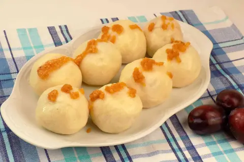 Pyszne knedle ze śliwkami, posypane okruszkami, na talerzu z niebiesko-białą kratką. Obok leżą świeże śliwki.