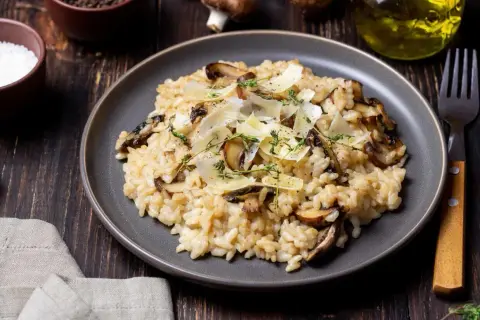 Aksamitne risotto z pieczarkami, posypane parmezanem i świeżym tymiankiem, prezentuje się apetycznie na ciemnym talerzu.
