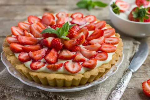 Pyszna tarta z truskawkami, udekorowana listkami mięty, na drewnianym blacie. Obok miseczka z deserem i nóż.