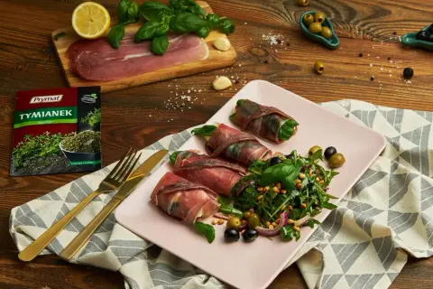 Roladki z szynką parmeńską i anchois, podane z rukolą, oliwkami i orzeszkami pinii. Obok talerza złote sztućce i opakowanie tymianku.