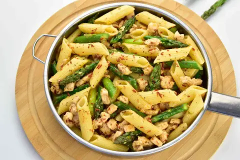 Penne z kurczakiem i szparagami, posypane płatkami chili, w metalowej misce na drewnianej desce.