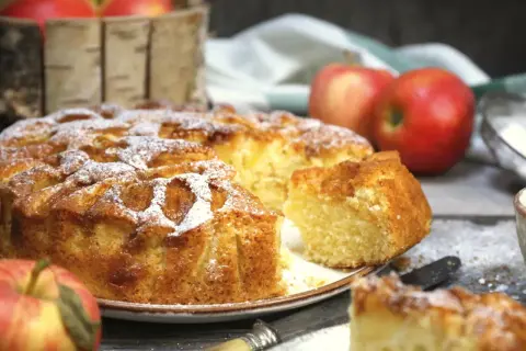 Puszyste ciasto na oleju z jabłkami, posypane cukrem pudrem. Obok jabłka i nóż.
