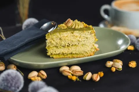 Pyszne ciasto z kremem pistacjowym, ozdobione orzechami i podane na talerzu. Obok filiżanka kawy i łyżeczka.