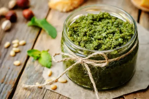 Słoik domowego pesto z bazylii, ozdobiony sznurkiem, na drewnianym blacie. Obok leżą orzeszki piniowe i listki bazylii.