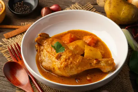 Pyszny kurczak w sosie curry z ziemniakami, podany w białej misce na lnianym tle. Obok widoczne przyprawy i drewniane sztućce.