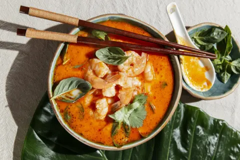 Aksamitna zupa tom yum z krewetkami, świeżą bazylią i kolendrą, podana z pałeczkami i łyżką.