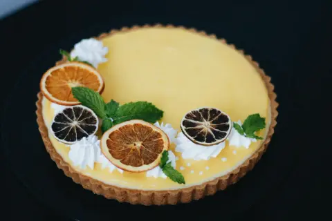 Pyszna tarta z owocami: cytrynowy krem, suszone pomarańcze i limonki, listki mięty i ozdobne kleksy bitej śmietany.