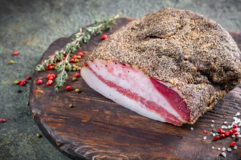 Guanciale co to? Włoski, peklowany policzek wieprzowy, pokryty pieprzem, leży na drewnianej desce z ziołami i pieprzem.