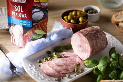 Pyszna szynka z szynkowara, pokrojona w plastry, podana z oliwkami, kaparami i świeżą bazylią. Obok opakowanie soli peklowej Prymat.