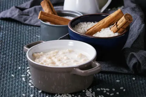 Gotowy do podania ryż na mleku w ceramicznej miseczce, obok miseczki z ryżem i laskami cynamonu.
