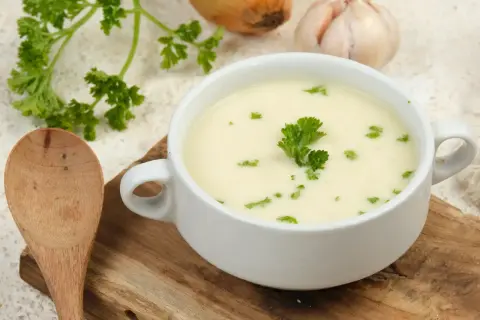 Aksamitna zupa krem z białych warzyw, udekorowana natką pietruszki, obok drewniana łyżka, cebula i czosnek.