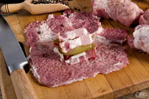 Roladki z karkówki w trakcie przygotowania. Na desce leżą rozbite plastry mięsa, doprawione pieprzem i solą, z dodatkiem boczku i ogórka kiszonego. Obok nóż i drewniana łyżka z ziarnami pieprzu.