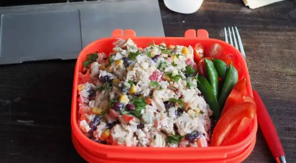 Pyszna sałatka z ryżem, kurczakiem, kukurydzą i fasolą w pomarańczowym pudełku. Obok pomidorki, groszek i papryka. Idealny lunch do pracy.