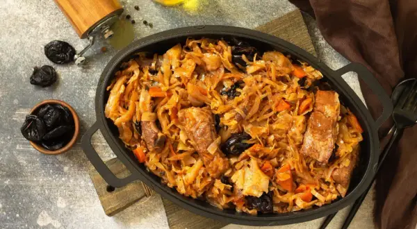 Pyszny bigos z kiszonej kapusty z mięsem i śliwkami, podany w żeliwnej patelni. Obok miseczka ze śliwkami i młynek do pieprzu.