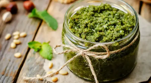 Słoik domowego pesto z bazylii, ozdobiony sznurkiem, na drewnianym blacie. Obok leżą orzeszki piniowe i listki bazylii.