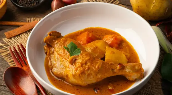 Pyszny kurczak w sosie curry z ziemniakami, podany w białej misce na lnianym tle. Obok widoczne przyprawy i drewniane sztućce.