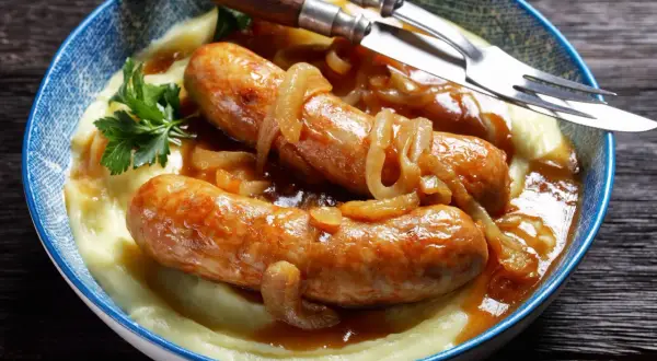 Złocista kiełbasa z cebulą i kremowym purée ziemniaczanym, udekorowana natką pietruszki. Danie podane w niebieskiej misce, gotowe do jedzenia.