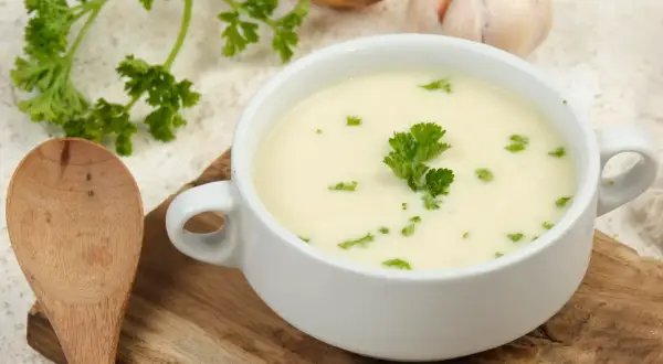 Aksamitna zupa krem z białych warzyw, udekorowana natką pietruszki, obok drewniana łyżka, cebula i czosnek.