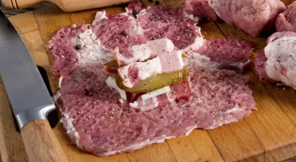 Roladki z karkówki w trakcie przygotowania. Na desce leżą rozbite plastry mięsa, doprawione pieprzem i solą, z dodatkiem boczku i ogórka kiszonego. Obok nóż i drewniana łyżka z ziarnami pieprzu.