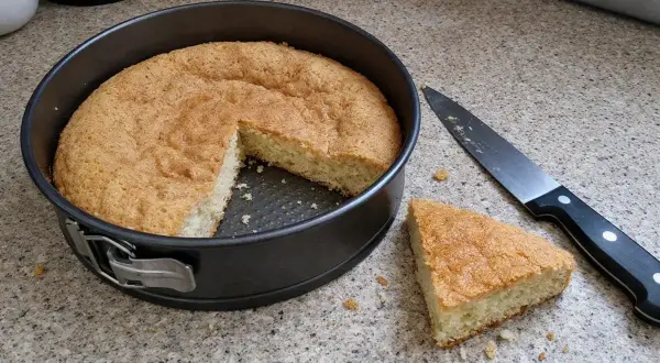 Puszysty biszkopt na tort w tortownicy, obok nóż i kawałek ciasta. Idealny do dalszego dekorowania.