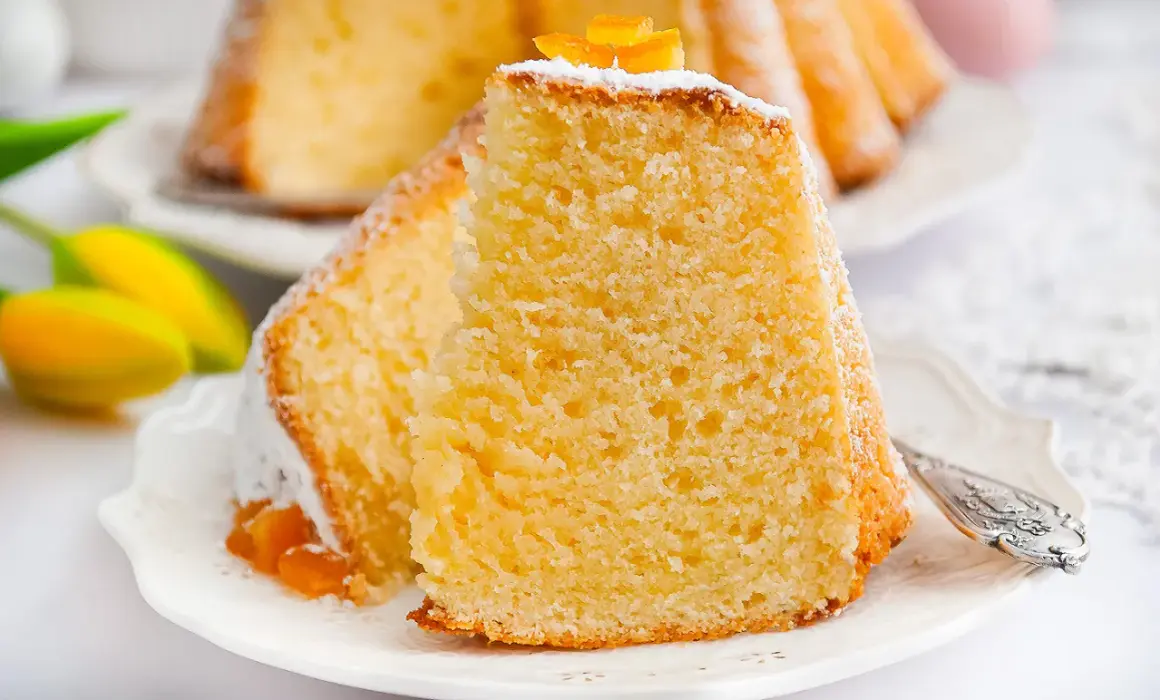 Puszysta babka z mascarpone, oprószona cukrem pudrem i ozdobiona skórką pomarańczową, na talerzyku z tulipanami.