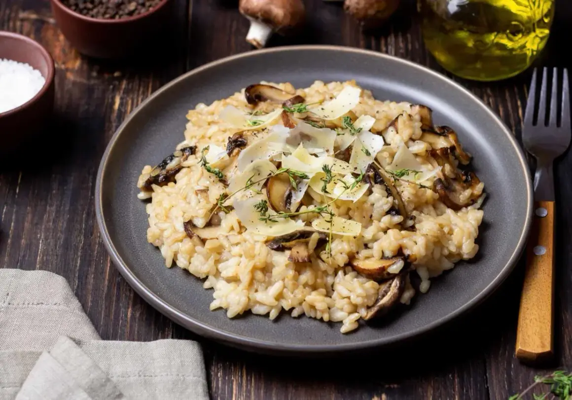 Aksamitne risotto z pieczarkami, posypane parmezanem i świeżym tymiankiem, prezentuje się apetycznie na ciemnym talerzu.