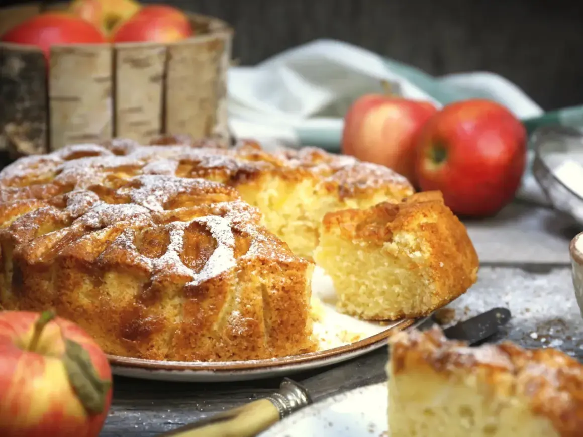 Puszyste ciasto na oleju z jabłkami, posypane cukrem pudrem. Obok jabłka i nóż.