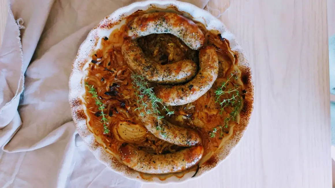 Złocista biała kiełbasa w piwie, zapiekana z cebulą i tymiankiem, prezentuje się apetycznie w ceramicznym naczyniu.