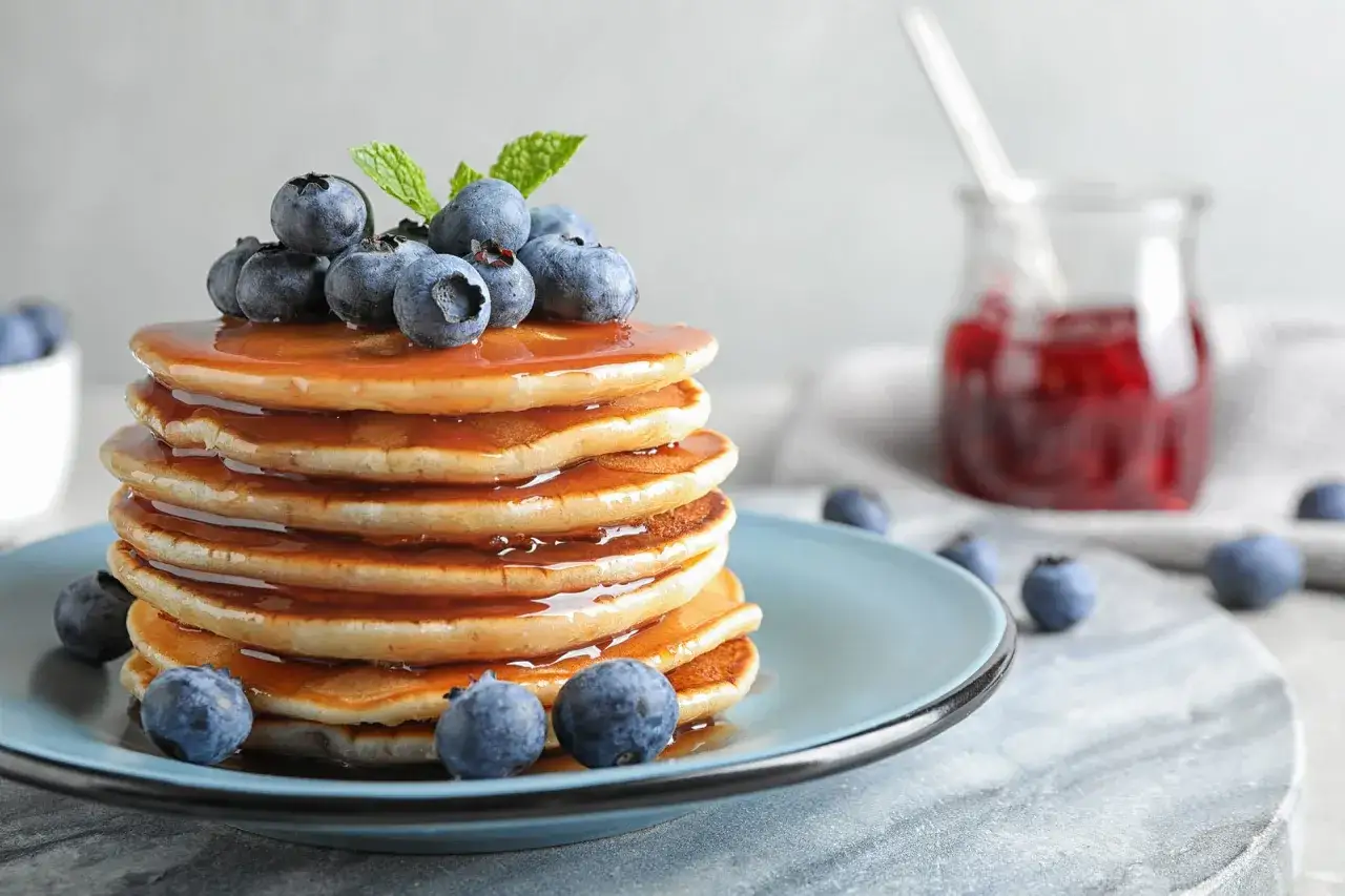 Idealny przepis na pancake: stos złotych placuszków polanych syropem, ozdobiony borówkami i miętą. Obok słoiczek z dżemem.